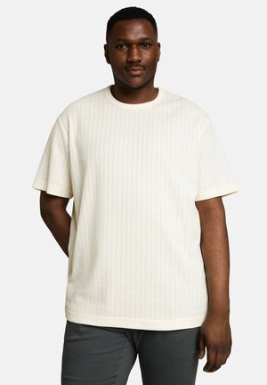Man with dark skin wearing a plain off-white short-sleeve shirt and dark pants standing against a light background.