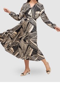 Robe longue et fluide en tissu léger. Présente un motif abstrait noir et crème, des manches longues et un décolleté en V. Associée à des talons crème.