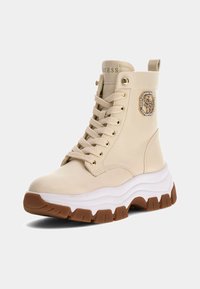Stivali da combattimento beige realizzati in pelle liscia, con punta rotonda, suola intermedia bianca spessa, suola esterna in gomma marrone e dettaglio del logo con cristalli.