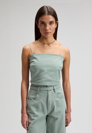 Femme de face portant un débardeur court vert clair à fines bretelles et un pantalon taille haute avec une ceinture assortie.