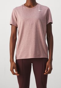 Osoba ubrana w koszulkę Nike o krótkim rękawie w heather mauve i ciemnoróżowe legginsy na jednolitym tle.