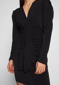 Svart klänning med långa ärmar, v-ringning och rynkdetaljer. Mjuk tyg med draperad accent i fronten. Slät textur.