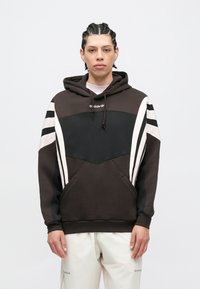 Fiatal férfi fonott hajjal, fekete-fehér Adidas kapucnis pulóvert és fehér nadrágot viselve, egyszínű világos háttér előtt áll.