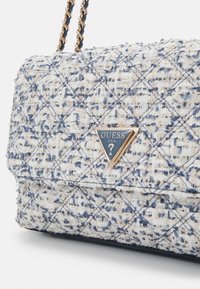 Sac à main en tissu tweed matelassé avec bandoulière chaîne dorée et plaque logo triangulaire Guess en bleu et or sur le rabat.