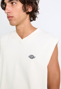 Hvit ermeløs strikket topp med v-hals, med en liten svart logo på brystet. Glatt tekstur og avslappet design.