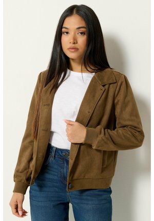 Junge Frau mit langen dunklen Haaren, die eine braune Jacke über einem weißen Hemd und blauen Jeans trägt, steht vor einem schlichten hellen Hintergrund.