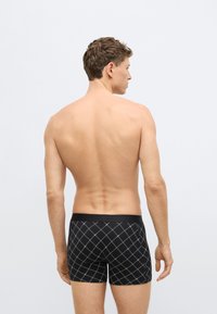 Crne bokserice s uzorkom dijamanta, elastičnim pojasom i kratkim nogavicama, koje nosi osoba prikazana sa stražnje strane.