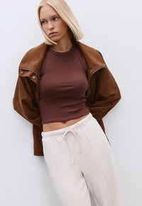 Braune Wildlederjacke über einem enganliegenden, dunkelbraunen Cropped-Top, kombiniert mit hellbeigen Jogginghosen mit Kordelzug und Seitennähten.