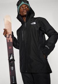 Schwarz wasserdichte Jacke mit Kapuze und Reißverschlüssen, mit "GORE-TEX" Branding. Das Model hält braune und schwarze Skier mit Bindungen.