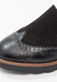 Chaussure en daim noir et cuir verni avec des détails brogués, un bout arrondi et une finition texturée ; elle présente une semelle marron avec une bordure contrastante.