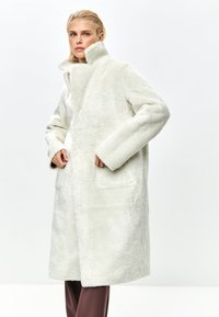 Weißer, strukturierten Pelzmantel mit hoher Kragen und vorderen Taschen. Das Design ist oversized, mit langen Ärmeln und gerader Silhouette.