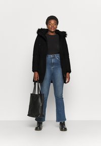 Parka preta com capuz de pelagem, cintura ajustável, combinada com jeans azuis de cintura alta e boca de sino e botins pretos. Bolsa tote preta na mão.