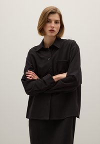 Camicia nera button-up realizzata in tessuto liscio. Presenta maniche lunghe, una tasca sul petto sinistro e una vestibilità rilassata con fondo dritto.