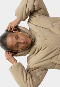 Beige pufferjakke med hætte, der har en quiltet design og knapper. Stoffet ser glat og let ud, med rummelige ærmer.