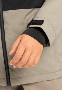 Zbliżenie na rękę blisko beżowego mankietu kurtki z czarnym wewnętrznym rękawem i czarnym zamkiem błyskawicznym na przedniej panele.