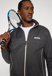 Μαύρο ελαφρύ μπουφάν με φερμουάρ στο μπροστινό μέρος, λογότυπο BOSS και ελαστικά μανίκια. Ελαφριά ρακέτα τένις κρατιέται στο άλλο χέρι του υποκειμένου.
