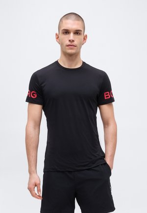 Young man with short hair wearing a black short-sleeve T-shirt with red letters on sleeves and black shorts, standing with one hand in pocket.