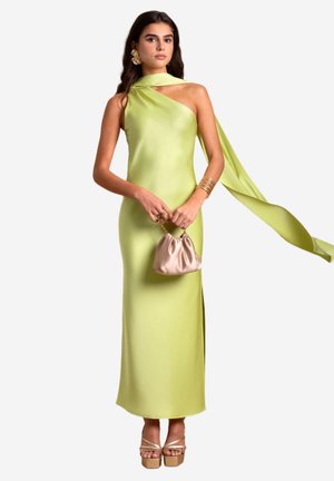 Robe asymétrique à une épaule en satin vert clair, dotée d'une fente sur le côté, accompagnée d'un sac en satin beige et de talons épais.