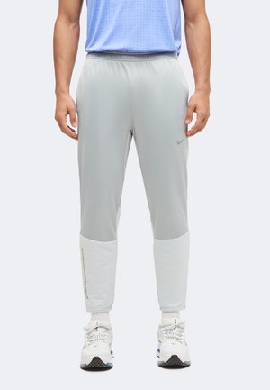 Homme portant un pantalon de sport gris clair à coupe fuselée avec des panneaux blancs au-dessus des chevilles, associé à des baskets blanches et une chemise bleu clair.