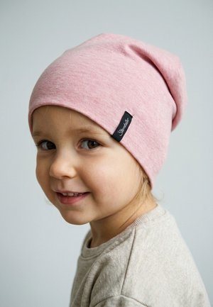 Young child smiling, wearing a light pink knit beanie with a black label on the side and a beige shirt, against a plain gray background.