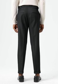 Pantalon noir sur mesure avec une texture lisse, coupe ajustée et ourlets retroussés, doté de deux poches arrière. Porté avec des chaussures habillées noires.