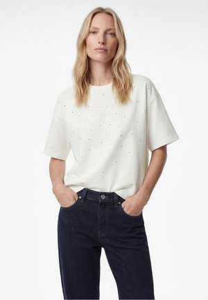 Blonde Frau mit offenem Haar, trägt ein weißes kurzärmeliges Hemd mit kleinen Punkten und dunkelblaue Jeans, Hände in den Taschen, steht vor weißem Hintergrund.