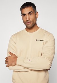 Beige Langarm-Sweatshirt mit geripptem Kragen und Bündchen; verfügt über ein gesticktes "Champion"-Logo auf der Brust und ein kleines Logo am Ärmel.