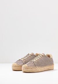 Des chaussures beige texturées avec une semelle en jute tissée, dotées d'œillets et de lacets dorés, et d'un accent de talon couleur crème.