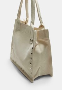 Vaaleanbeige kangaskassi, jossa on kaksi tukevahkoa kahvaa, tähdenmuotoiset niitit toisella sivulla ja vetoketjullinen yläosa.