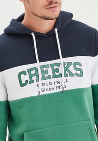 Felpa con cappuccio con blocchi di colore blu navy, bianco e verde. Presenta la scritta "CREEKS ORIGINAL Dal 1974", stringhe e una tasca frontale.
