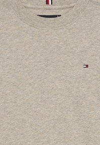 Lys grå genser laget av mykt stoff, har rund hals og en liten brodert logo med røde, hvite og blå detaljer på brystet.