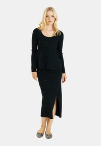 Vestido de suéter negro con cintura peplum y textura acanalada; presenta acentos de cuentas plateadas y una abertura lateral, combinado con zapatos planos adornados.