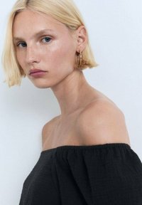 Schwarzes Off-Shoulder-Oberteil aus texturiertem Stoff mit lockerer Passform. Das Modell hat kurze blonde Haare und trägt Hängohrringe, vor einem hellen Hintergrund.