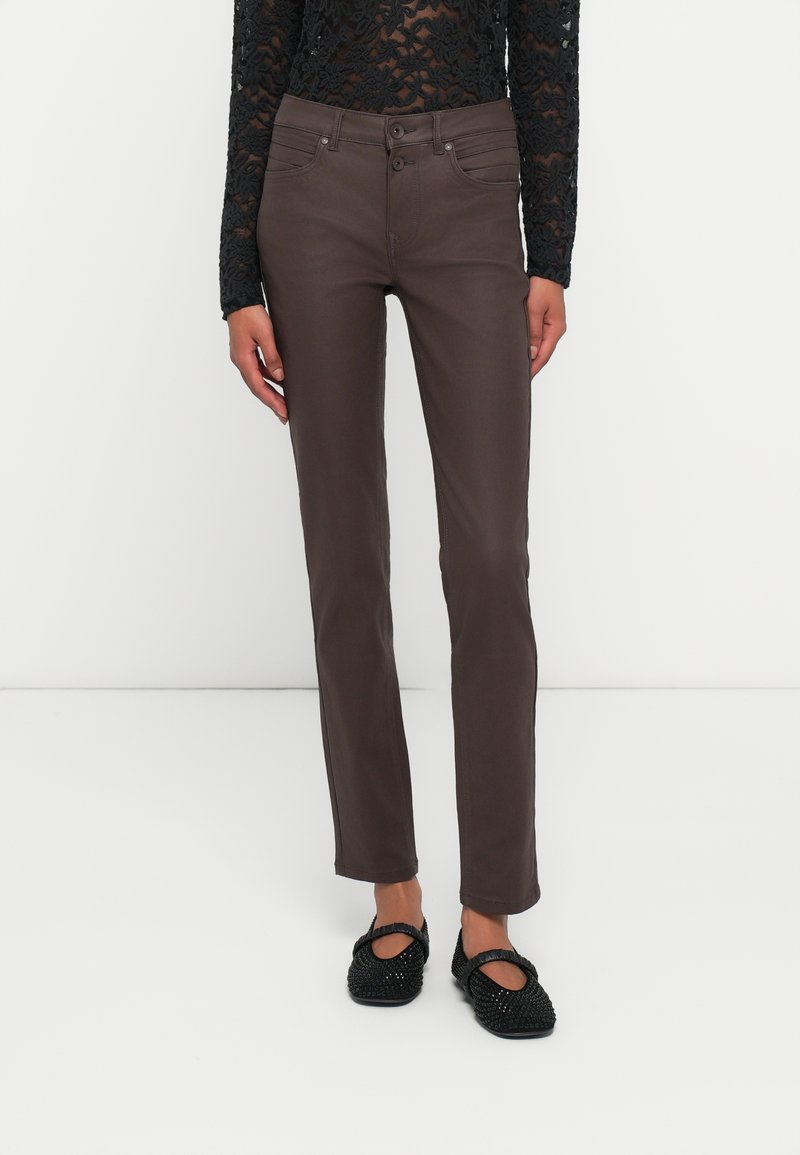 Femeie îmbrăcată cu pantaloni slim-fit maro închis, bluză neagră cu dantelă și mâneci lungi, și pantofi tip Mary Jane cu textură neagră, stând pe un fundal alb.