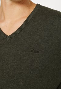 Nahaufnahme der oberen Brust einer Person, die einen dunkelgrünen V-Ausschnitt-Pullover mit einem dezenten gestickten Logo auf der linken Seite trägt.