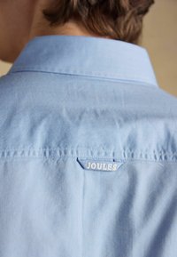 Vue arrière d'une personne portant une chemise à col bleu clair avec une étiquette "JOULES" cousue sous le col, dans le haut du dos.