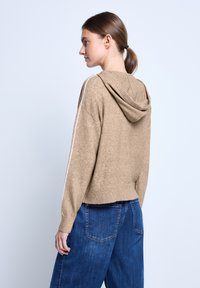 Brauner Kapuzenpullover mit einer weichen Textur und cropped Design, ausgestattet mit weißen Streifen an den Ärmeln und einem gerippten Saum. Kombiniert mit blauen Jeans.