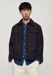 Schwarze, kurzärmlige Jacke mit Kragen, zwei Brusttaschen und entspannter Passform, getragen über einem blauen Denim-Hemd; glatte Textur, minimales Design.