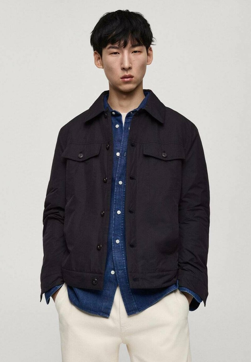 Schwarze, kurzärmlige Jacke mit Kragen, zwei Brusttaschen und entspannter Passform, getragen über einem blauen Denim-Hemd; glatte Textur, minimales Design.