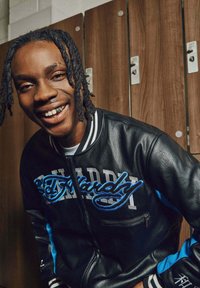 Young man with braided hair wearing a black and blue Ed Hardy jacket smiles in front of wooden lockers.