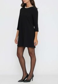 Schwarzes Shiftkleid mit Dreiviertelärmeln und einem metallischen Akzent am Ausschnitt, kombiniert mit schwarzen High-Heels und transparenten schwarzen Strumpfhosen.