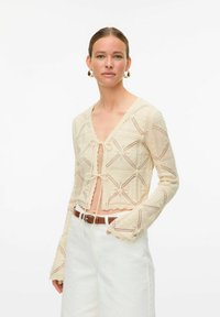 Gebreide cardigan in crème met een textuurpatroon in geometrische vorm, striksluiting en trompetmouwen, gecombineerd met witte high-waisted broeken.