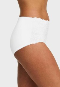 Vrouw in witte hoog uitgesneden onderbroek met kantdetails aan de zijkanten, getoond van de taille tot halverwege het bovenbeen tegen een effen achtergrond.