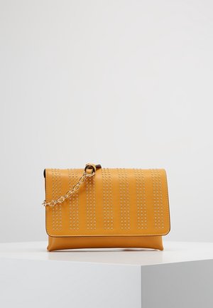Mosterdgele crossbodytas met verticale zilveren studs en een gouden kettingriem, geplaatst op een wit displaypodium tegen een effen achtergrond.