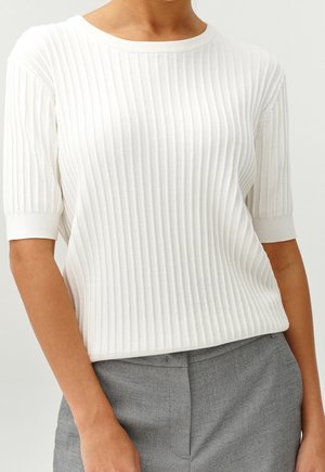 Woman wearing a white ribbed short-sleeve knit top and high-waisted gray tailored pants, standing against a light background.