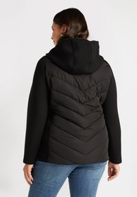 Svart pufferväst med en texturerad chevron-design på baksidan, med huva och långa ärmar, tillsammans med blå jeans.