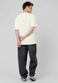 T-shirt blanc texturé à manches courtes avec une coupe décontractée, associé à un jean baggy gris foncé. La personne se tient avec les mains jointes derrière le dos.