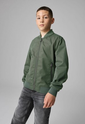 Zielona kurtka bomberka z gładkiego materiału, z zamkiem z przodu, ściągaczami na rękawach i haftowanym logo. W zestawie z ciemnoszarymi dżinsami.