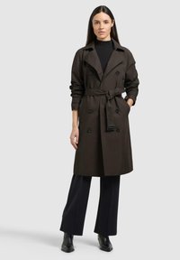 Donna che indossa un trench marrone scuro con cintura sopra un dolcevita nero e pantaloni a gamba larga, in piedi con una mano in tasca contro uno sfondo semplice.