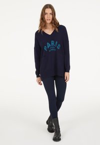 Navy oversized sweater with "PARIS MON AMOUR" in sequined turquoise. Features v-neck, two front pockets, and ribbed hem. Worn with dark leggings and boots.